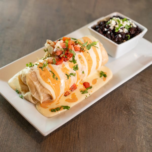 A Dahlia burrito covered in white and orange sauce, topped with pico de gallo, served with black beans.