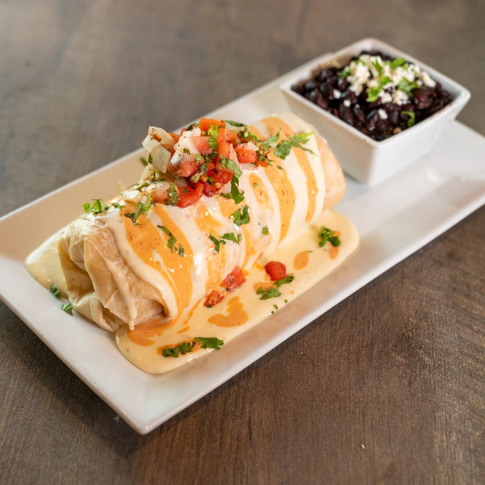 A Dahlia burrito covered in white and orange sauce, topped with pico de gallo, served with black beans.