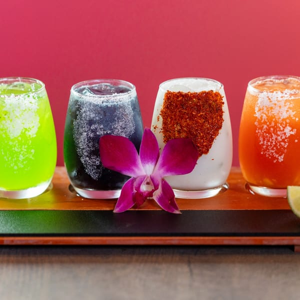 Four colorful Dahlia margaritas, including green, black, white with chili, and orange, on a wooden tray.
