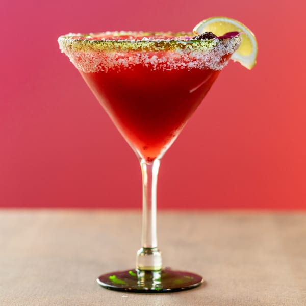 A vibrant red Dahlia margarita in a martini glass, garnished with lime and a salt rim.